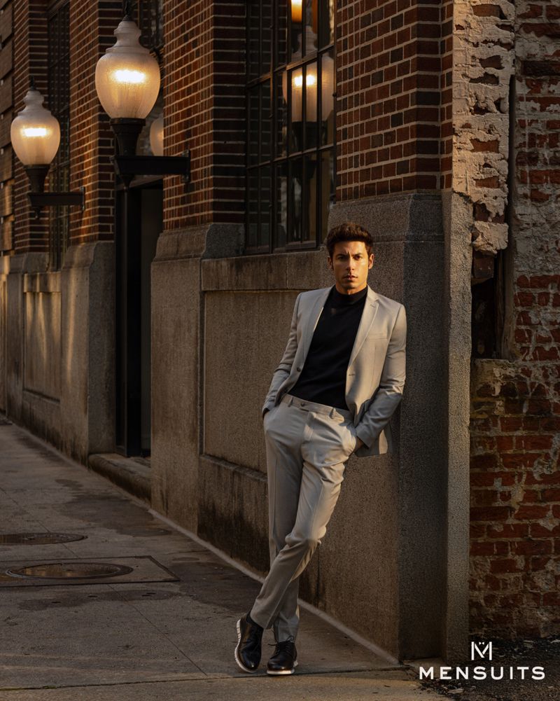 An adult male model leans against a brick wall in a grey suit, illuminated by streetlights in an urban outdoor setting. 