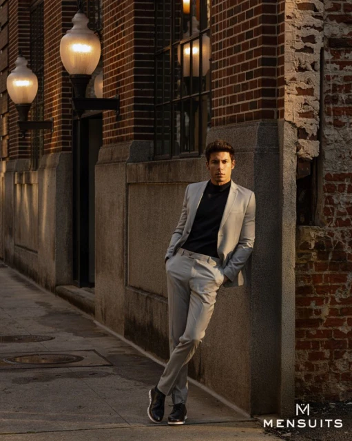 An adult male model leans against a brick wall in a grey suit, illuminated by streetlights in an urban outdoor setting. 