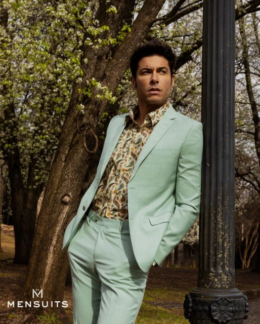 Adult male model leans against a light post outdoors, wearing a mint green suit and patterned shirt, showcasing menswear fashion.
