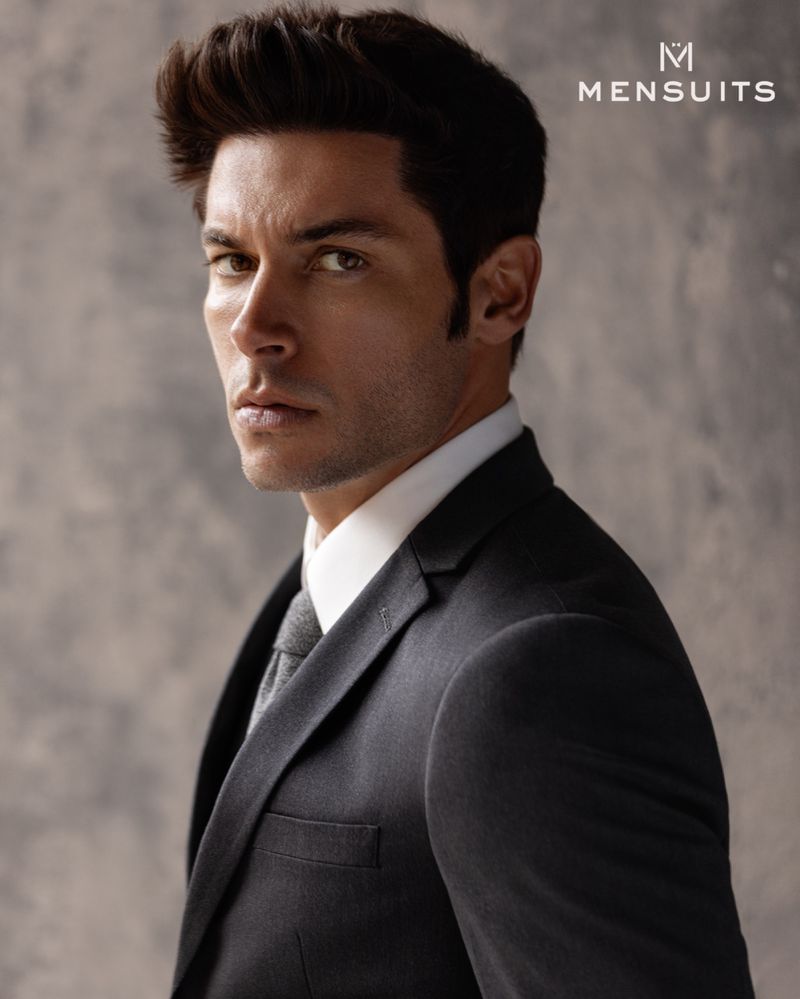 Portrait of adult male in dark suit and grey tie looking to camera against muted grey textured background in studio-style lighting.