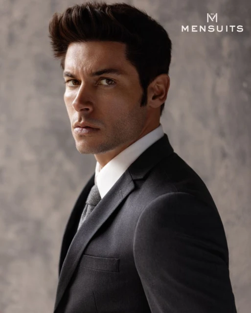 Portrait of adult male in dark suit and grey tie looking to camera against muted grey textured background in studio-style lighting.