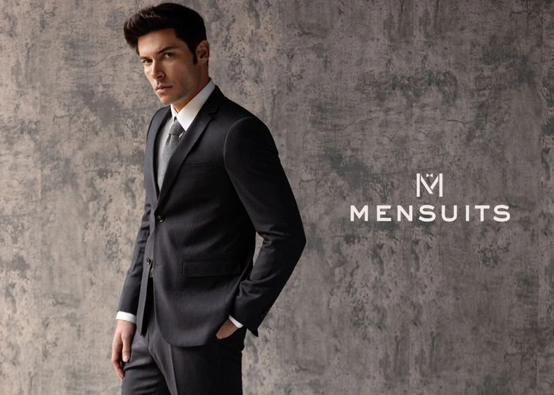 Portrait of a handsome adult male fashion model posing in a dark suit, white shirt, and tie against a grey textured backdrop indoors.