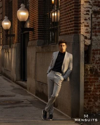 Man wearing a light grey suit and black shirt leaning against a brick building on a city street at dusk.