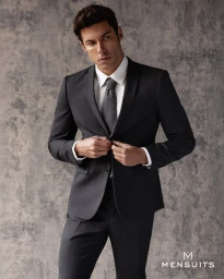 Model Andrea Denver in a dark charcoal suit adjusting his jacket against a textured concrete wall.