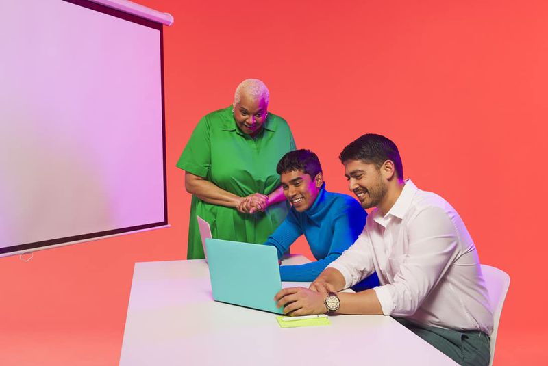 Three people gather around a laptop at a white table in a coral-colored room, with a screen behind them. They are smiling.