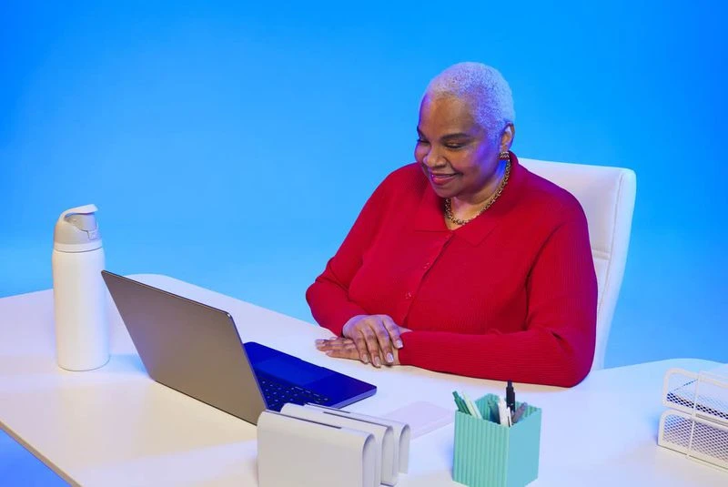 Mature woman in a red sweater works at her desk on a laptop against a blue background. Water bottle and office supplies nearby.