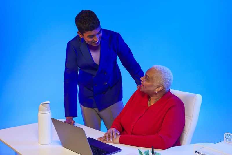 Young adult in a suit looks on at an older woman sitting at a desk with a laptop, set against a vibrant blue background.