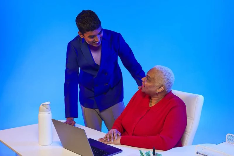 Young adult in a suit looks on at an older woman sitting at a desk with a laptop, set against a vibrant blue background.