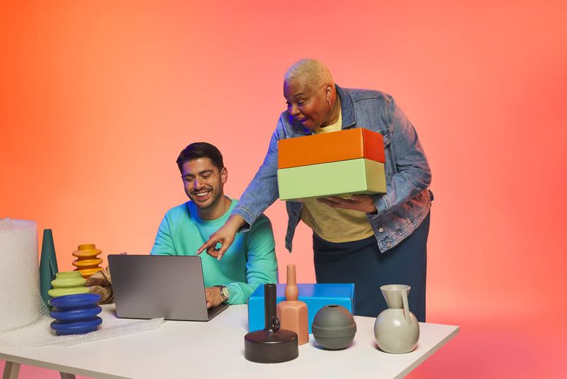 Two people are in a studio setting. One is pointing to a laptop, and the other holds a box. Vases and packing materials are visible.