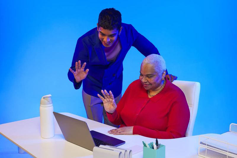 Two people, an adult male and an elderly female, wave during a video call. The pair sits at a desk in front of a blue background.