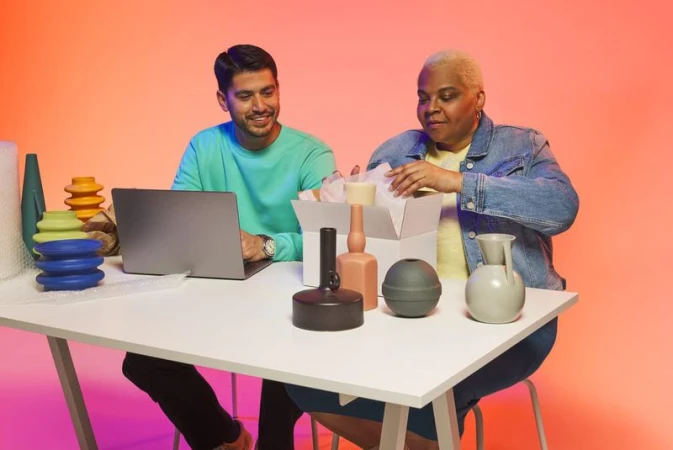 Two people sitting at a desk with a laptop and vases. The woman is holding a vase from a box, and the man is looking at the laptop.