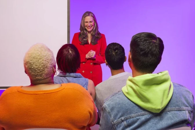 A smiling woman in a red suit stands presenting to a small group of diverse adults in an indoor setting against a lavender background.