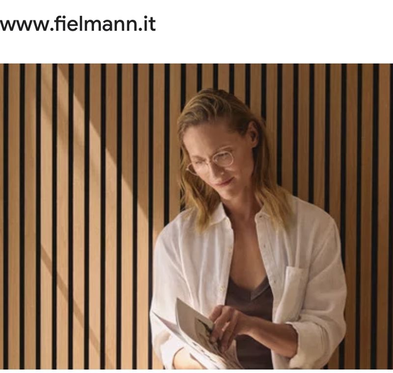 A woman with blonde hair and glasses is reading a magazine in front of a slatted wooden wall, wearing a white button-down shirt.
