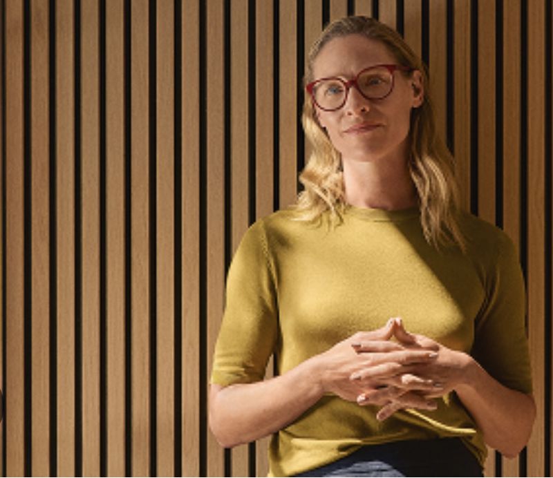 Indoor portrait of an adult woman with blonde hair wearing glasses and a yellow sweater standing in front of a vertical slat wall.