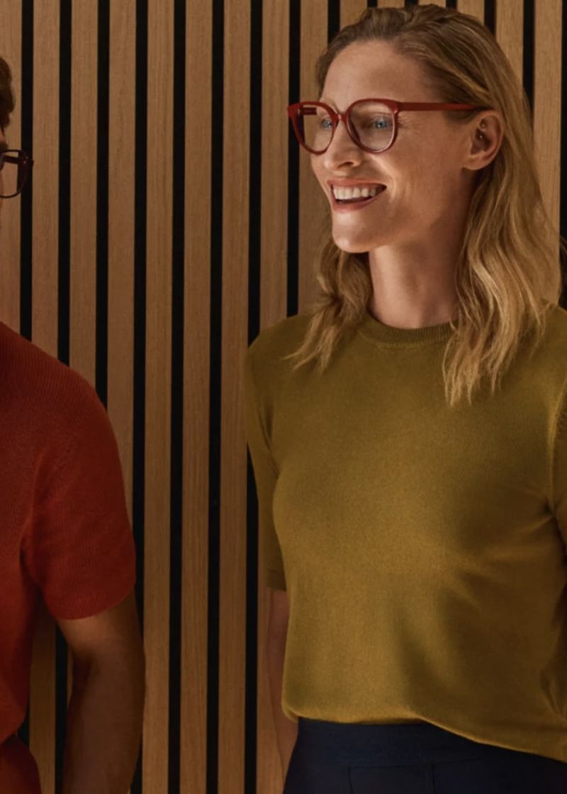 Woman with glasses smiles next to a man in a casual indoor setting with a vertically striped background, showcasing a natural, friendly interaction.