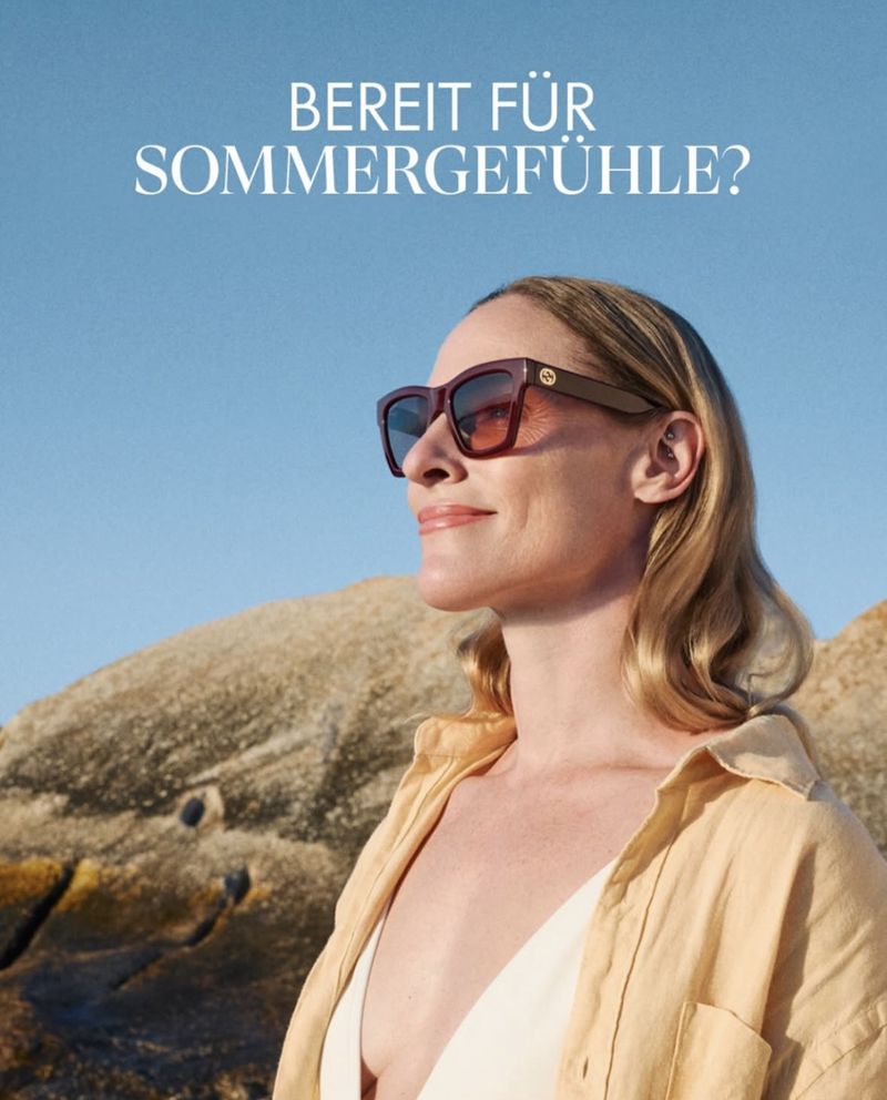 Woman in sunglasses and a yellow shirt posing on a beach with rock formations under a clear blue sky in a medium shot.