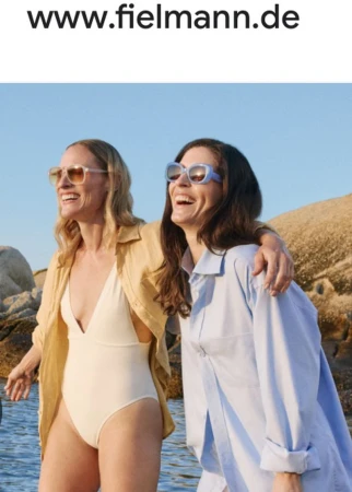 Two adult females smiling and posing on the beach with eyeglasses, wearing a swimsuit and a blue shirt, on a sunny day.