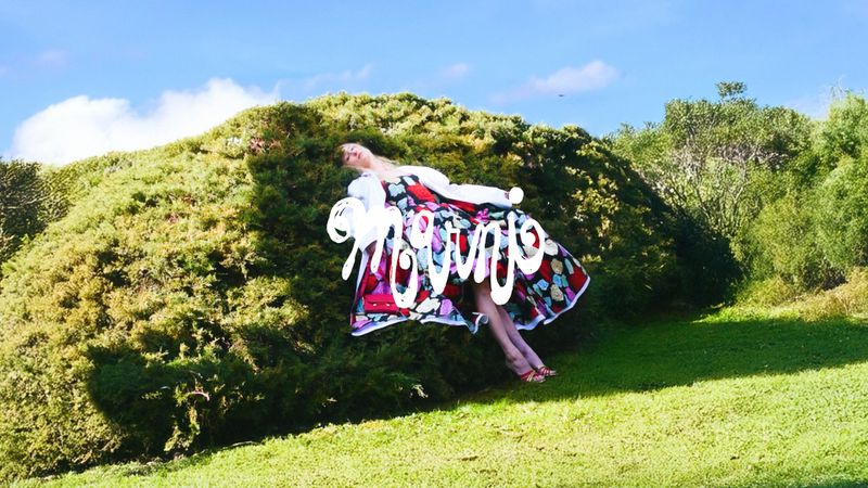 Woman in colourful dress lying on a grassy hill under a blue sky with artistic text overlay. Outdoors fashion shot in nature.
