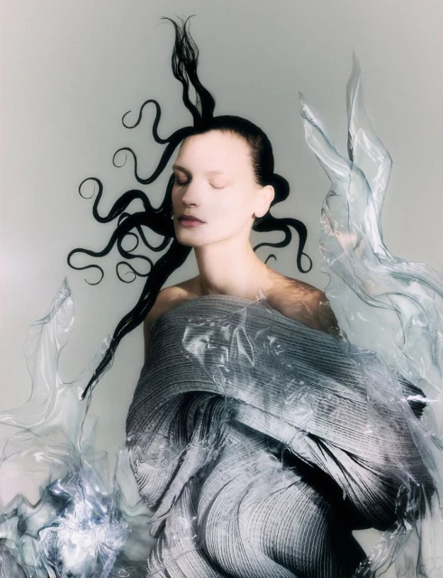 Woman with slicked-back dark hair, closed eyes, and gray sculpted draped fabric with shiny transparent plastic, set against a bright background.