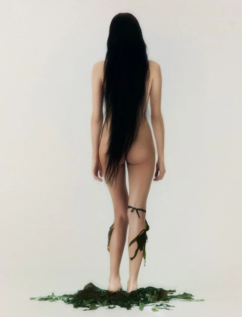 Back view of a nude woman with long black hair and dark green seaweed accents against a solid white backdrop in a studio setting.