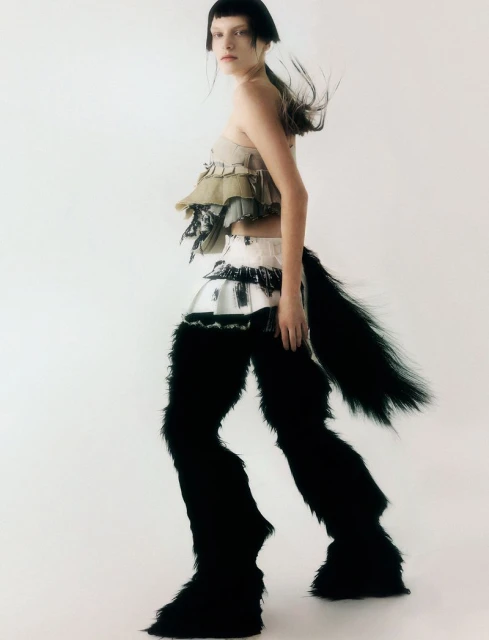 A woman poses in a light studio wearing a beige top and furry black pants with a tail, against a white backdrop.