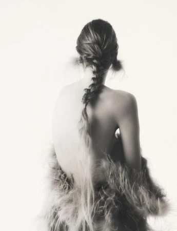 Back view of a woman with long braided hair, wearing a feathered shawl, in a studio setting with soft, diffused lighting.
