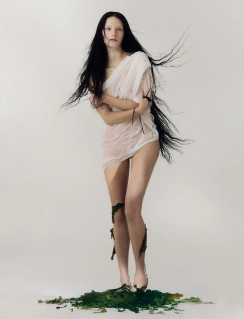 Young woman standing on greenery in translucent draped garment. She has long, dark hair and is posing in a photo studio setting.