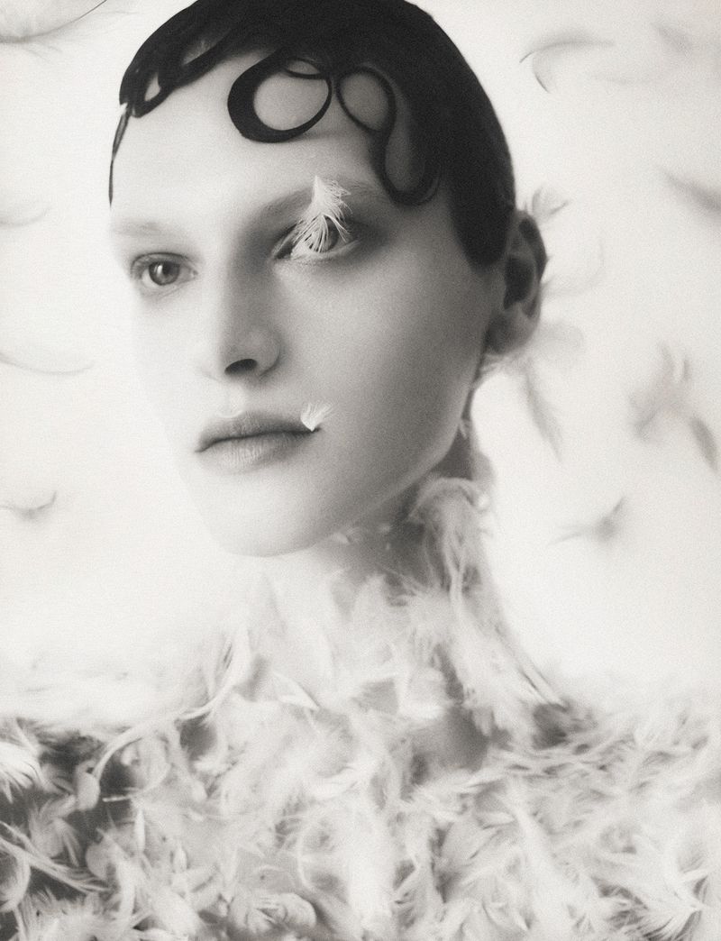 Monochrome portrait of a fair-skinned young woman adorned with feathers, against a soft white background in a studio fashion setting.