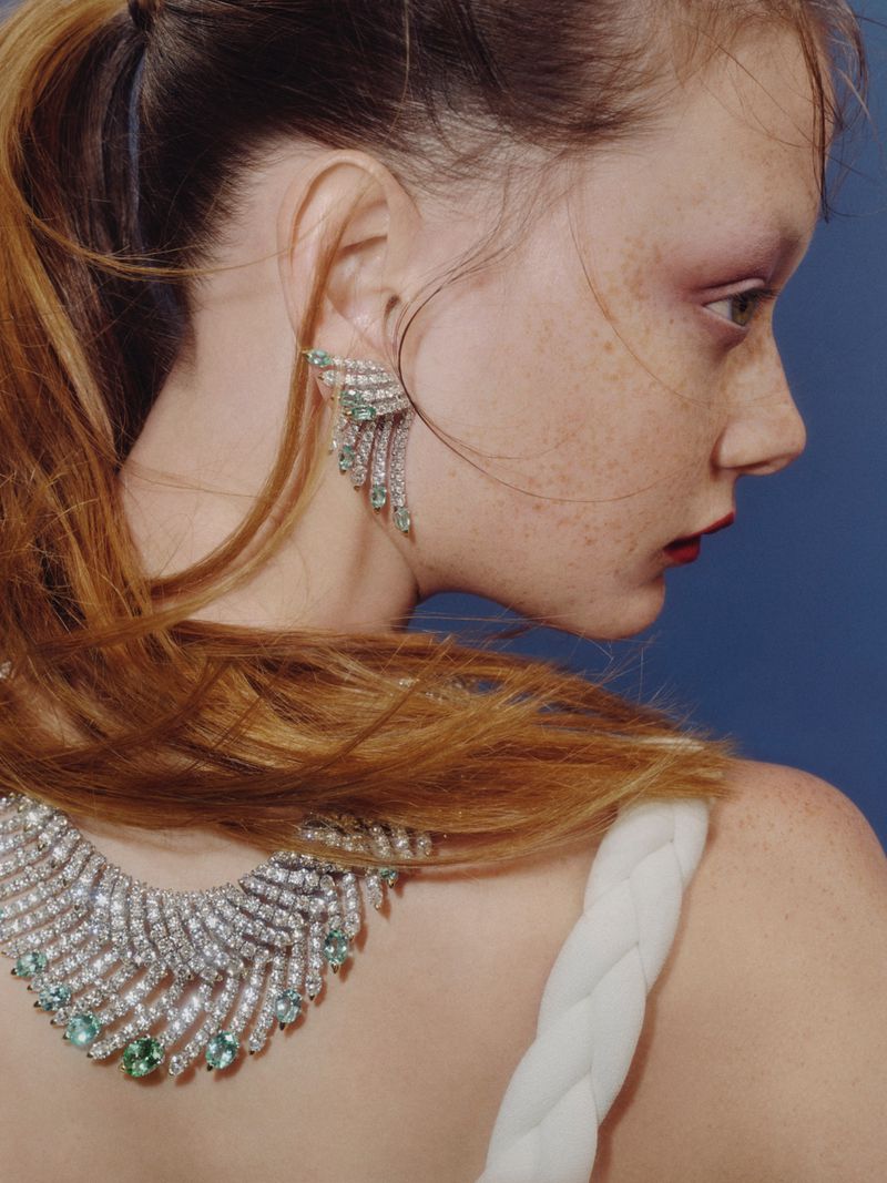 Elegant jewelry on a fair-skinned adult female with red hair pulled back, showcasing a necklace and earrings against a blue background.