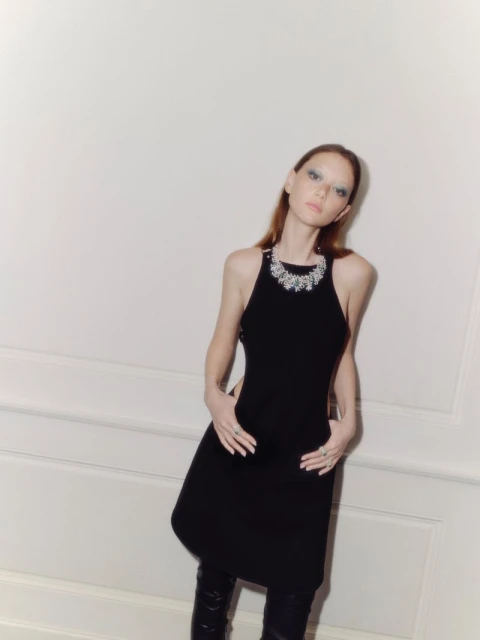 Fashion portrait of a stylish adult female in a black dress and high boots posing in front of a light wall with slight shadow.