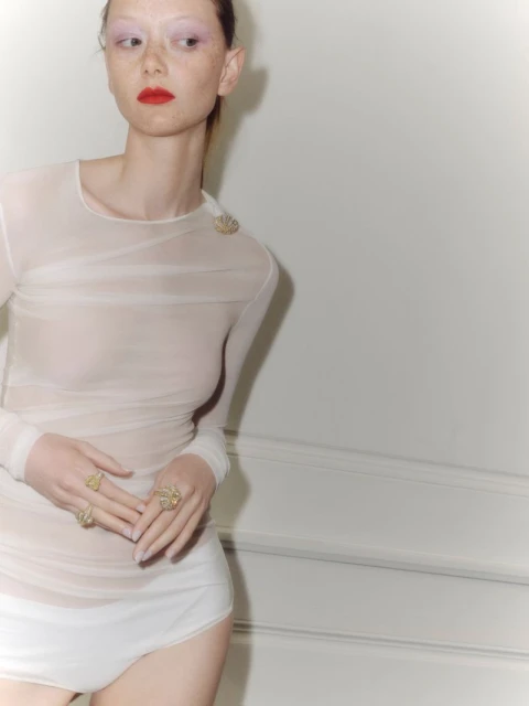 A woman poses indoors in a sheer white top and bottom with large ring jewelry and red lipstick looking right against a white wall.