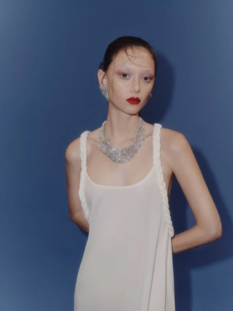 An adult female fashion model poses in a white dress with jewelry against a blue background in a medium studio shot. She has dark red lips.