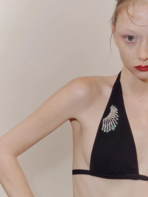 Close-up of a young woman with red lips and fair skin wearing a black bikini top adorned with a jeweled embellishment on a beige background.