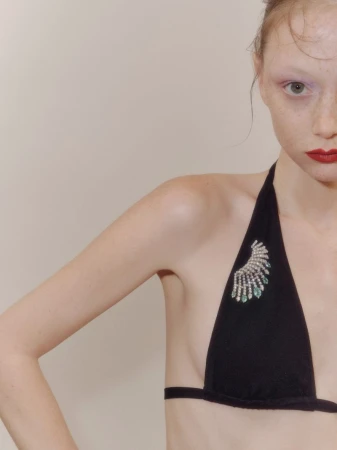 Close-up of a young woman with red lips and fair skin wearing a black bikini top adorned with a jeweled embellishment on a beige background.