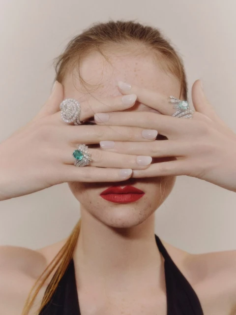 Model showcases gemstone rings covering eyes in studio setting. Jewellery fashion shot features three elegant statement rings on fingers.
