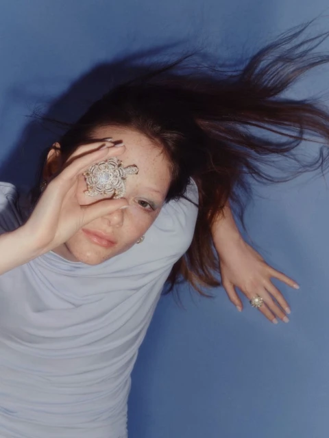 Portrait of a young woman with long brown hair holding a large ring in front of one eye, wearing a light purple top, against blue background.