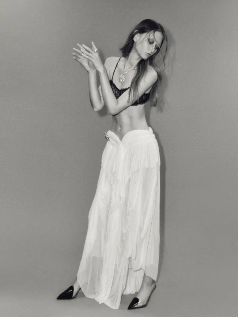 Monochrome portrait of a slender female fashion model in a black bralette, long white sheer skirt, and black heels posing in a photo studio.