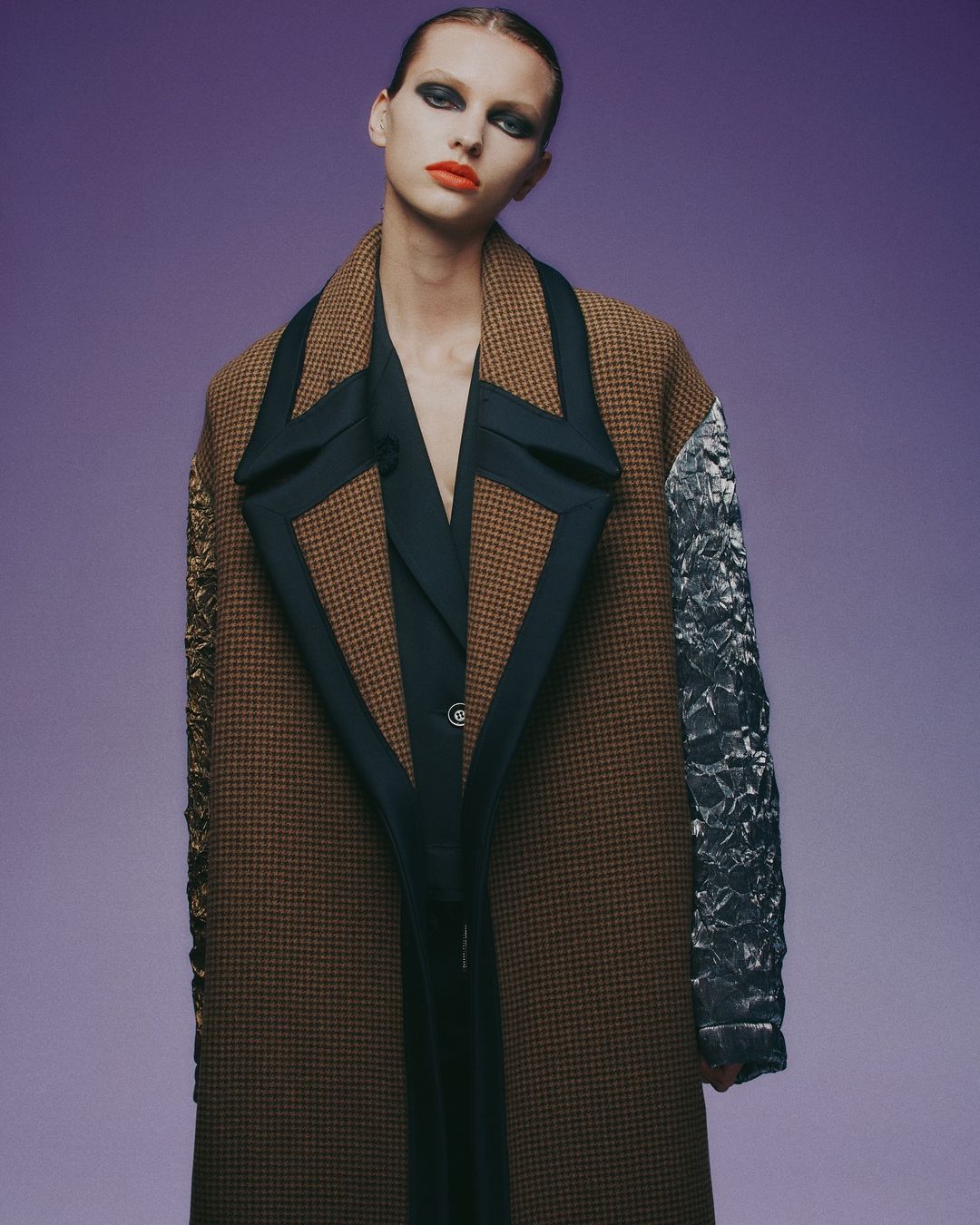 A woman in a stylish, oversized patterned coat poses in front of a violet backdrop, showcasing a high-fashion look and bold makeup.