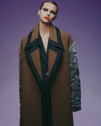 Model wearing a brown patterned coat with contrasting black lapels and metallic sleeves against a purple background.