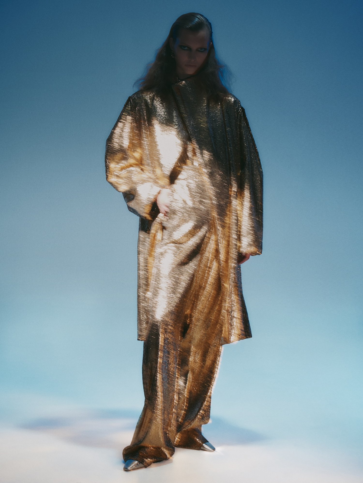 A fashion model poses in a full-length shiny gold lamé pantsuit with matching long coat against a light blue gradient background.