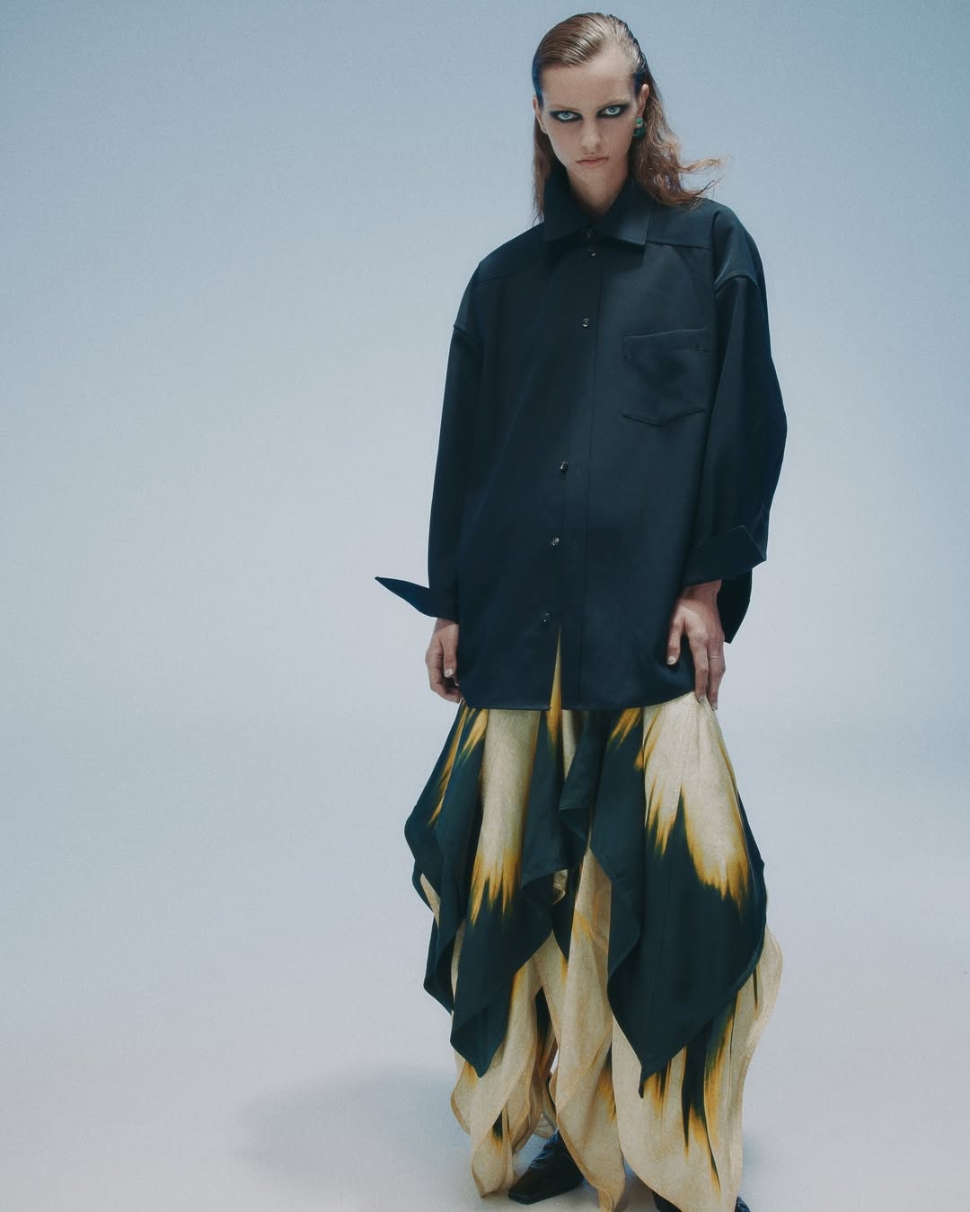 A woman poses in a black button-down shirt and a long patterned skirt against a pale blue background in a fashion forward studio portrait.