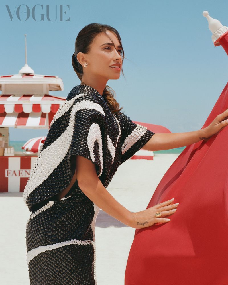 A tanned-skinned woman wearing a black and white patterned dress poses on a beach with a red lifeguard tower during daytime.