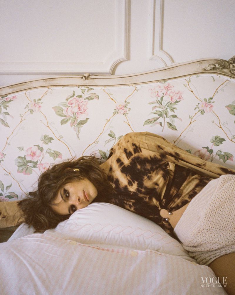 Brunette woman with brown hair and tie-dye top poses on a floral sofa in a studio; soft indoor light and a medium shot highlight her look.
