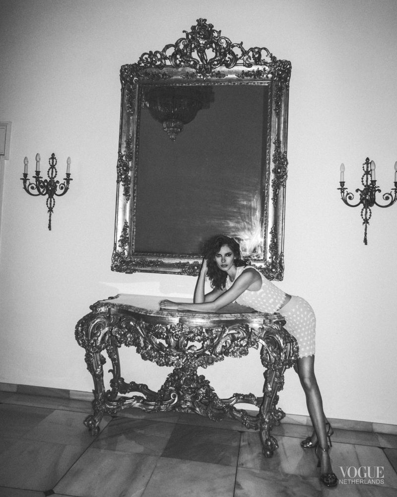 A vintage-style portrait captures a woman posing on an ornate console table, adorned with a large mirror, set against a bright background.