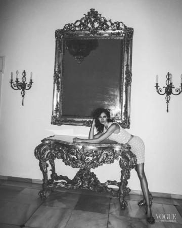 A vintage-style portrait captures a woman posing on an ornate console table, adorned with a large mirror, set against a bright background.
