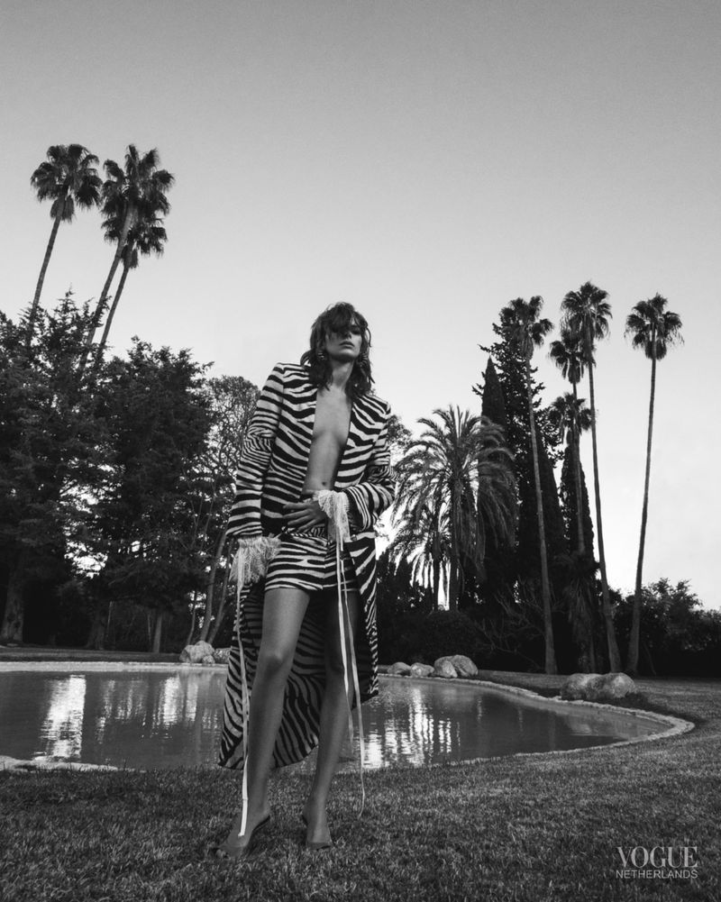 Fashion shot in black and white featuring a model in zebra-print attire standing on a lawn with palm trees and a pond.