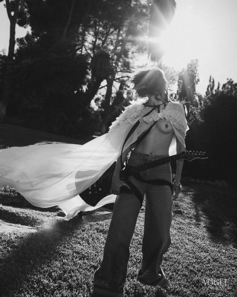 A woman stands outdoors in a field wearing jeans, a cape, and holding an electric guitar in a black-and-white fashion shot.