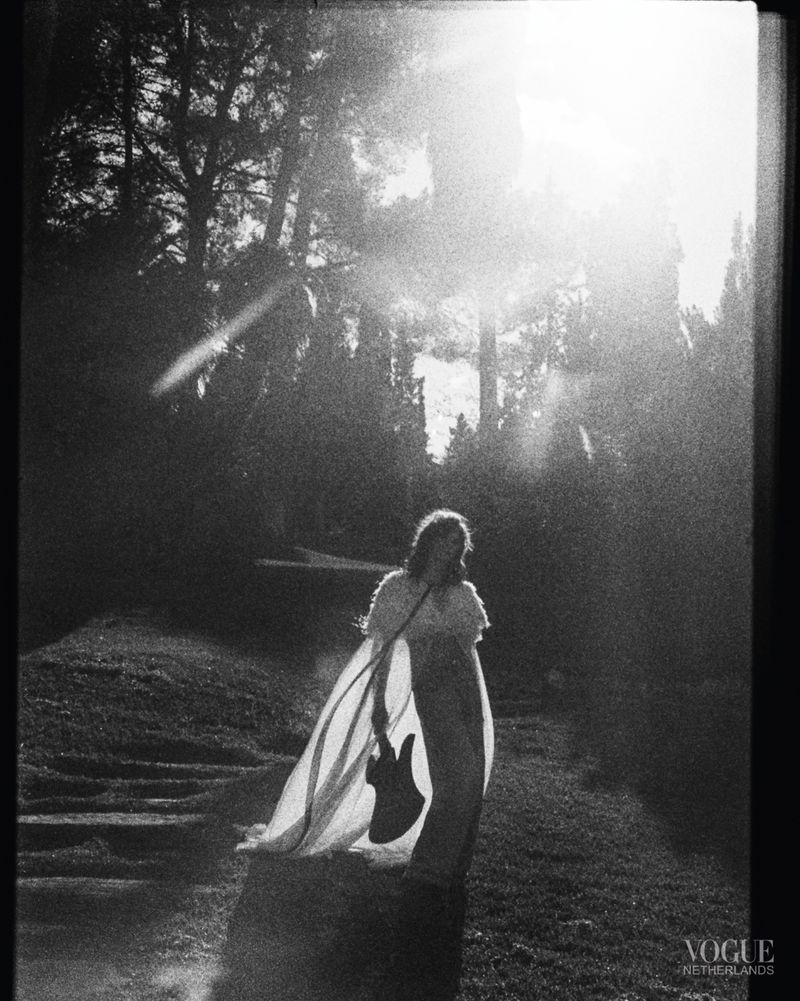 Monochrome shot of a woman standing in a wooded area, holding a guitar and wearing a flowing gown, sunlight streams through trees.