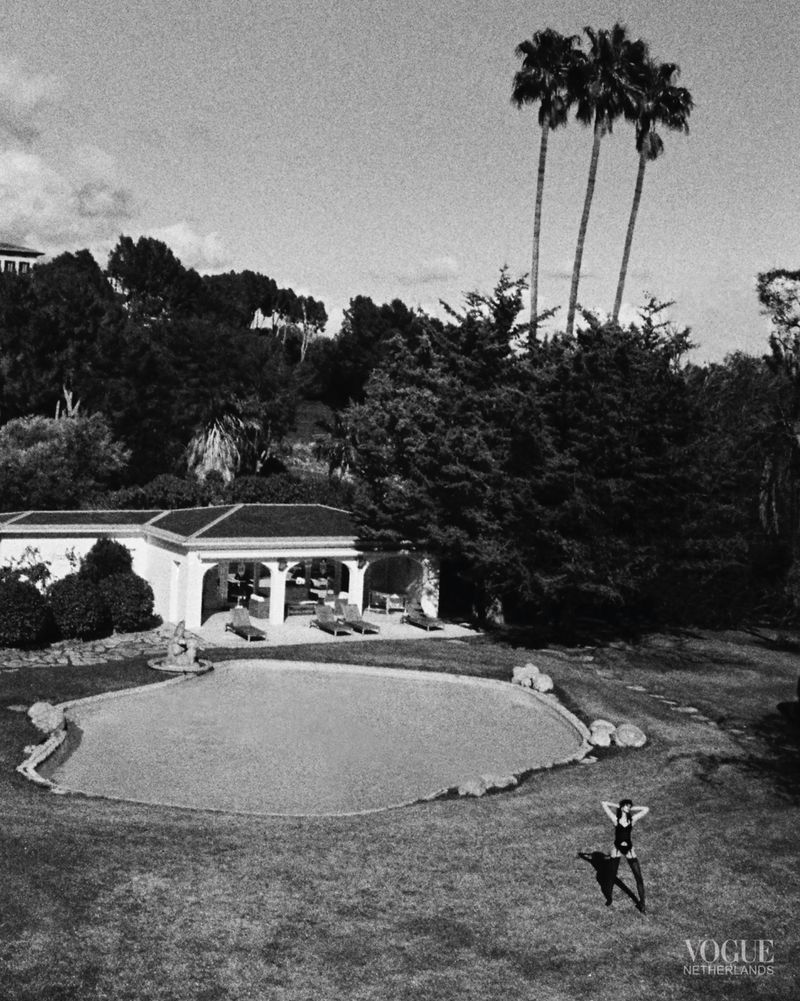 Monochrome shot of a woman in a swimsuit standing on green grass near a pool, trees, and a building on a sunny day.