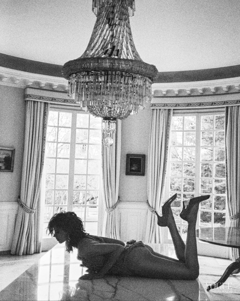 Monochrome photograph of a woman lying on a marble floor in a room with large windows and a chandelier, wearing heels.